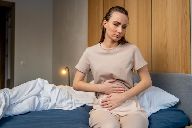 Una giovane donna con un'espressione facciale dolorosa che tiene il suo stomaco che soffre di dolori mestruali è seduta su un letto di casa con crampi allo stomaco