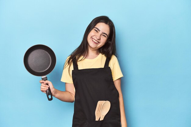Young woman with pan on blue studio happy smiling and cheerful