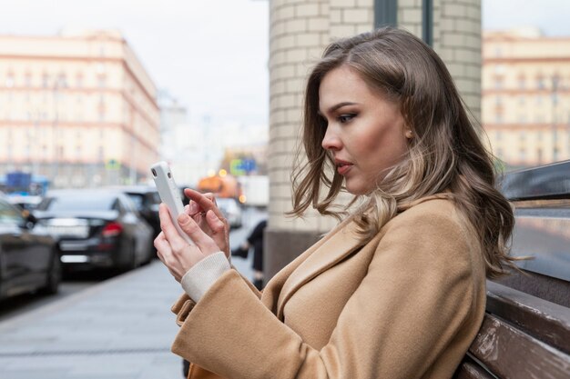 携帯電話を手にした若い女性が街のベンチに座っています。ベージュのコートを着た美しい金髪がメッセージを書いています。