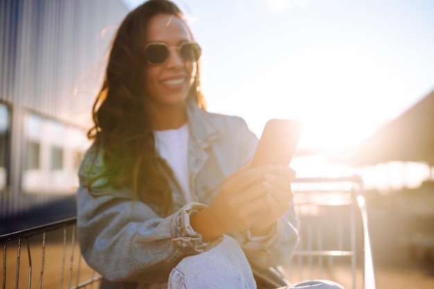 Young woman with phone sitting shopping cart having fun near color wall Blogging