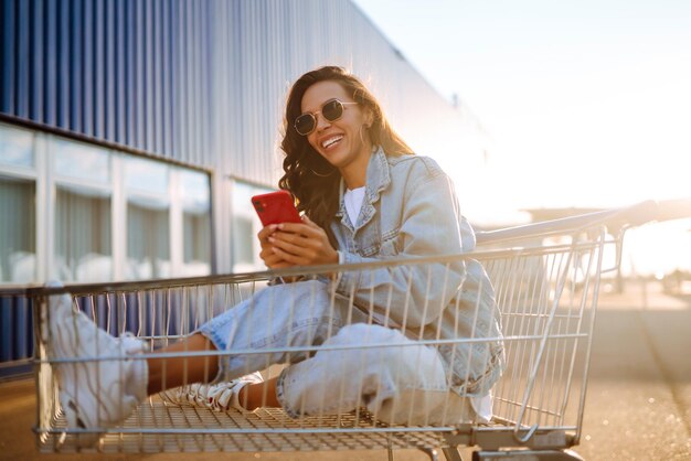 Young woman with phone sitting shopping cart having fun near color wall Blogging