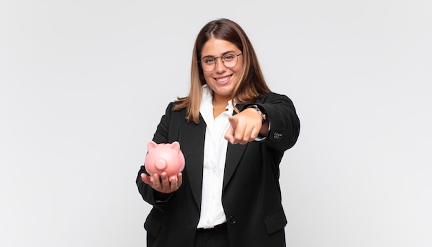 Young woman with a piggy bank pointing at front with a satisfied, confident, friendly smile, choosing you