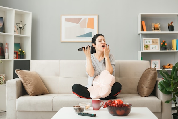 Young woman with pillow holding phone sitting on sofa behind coffee table in living room