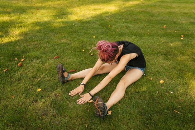 写真 日没時に公園で屋外でヨガの練習をしているピンクのドレッドヘアを持つ若い女性。初心者のハードワーク。