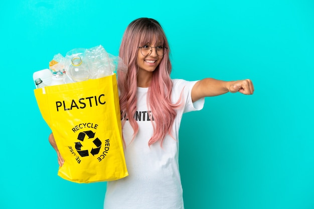 Молодая женщина с розовыми волосами держит сумку, полную пластиковых бутылок для переработки, изолированную на синем фоне, показывая жест рукой вверх