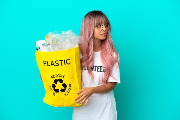 Young woman with pink hair holding a bag full of plastic bottles to recycle isolated on blue background looking to the side