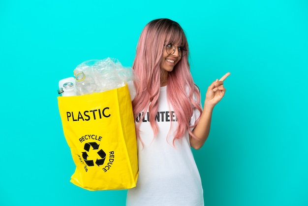 Foto giovane donna con i capelli rosa in possesso di un sacchetto pieno di bottiglie di plastica da riciclare isolato su sfondo blu che indica una grande idea