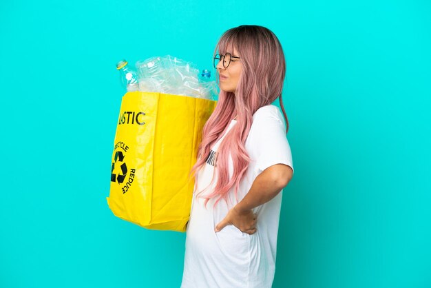 Молодая женщина с розовыми волосами, держащая мешок, полный пластиковых бутылок для переработки, изолирована на синем фоне и страдает от боли в спине из-за того, что приложила усилия