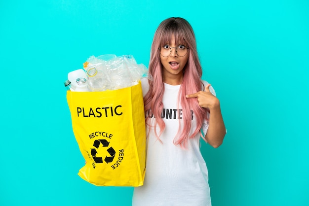 Young woman with pink hair holding a bag full of plastic bottles to recycle isolated on blue background with surprise facial expression