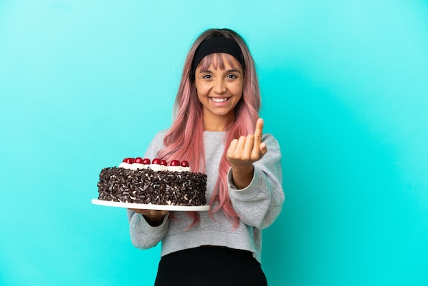 Молодая женщина с розовыми волосами держит праздничный торт на синем фоне, делая приближающийся жест