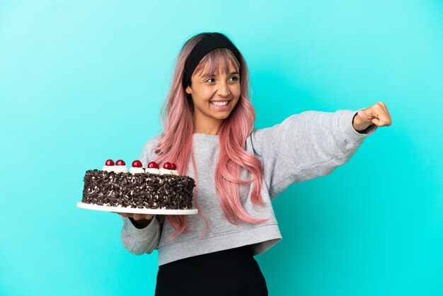 Young woman with pink hair holding birthday cake isolated on blue background giving a thumbs up gesture