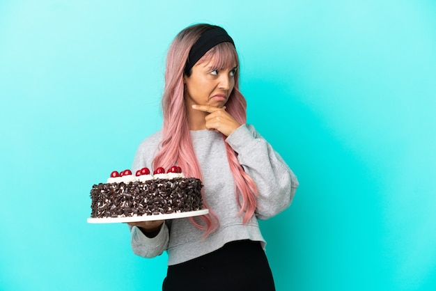 Young woman with pink hair holding birthday cake isolated on blue background having doubts