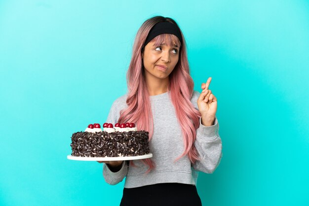 Young woman with pink hair holding birthday cake isolated on blue background with fingers crossing and wishing the best