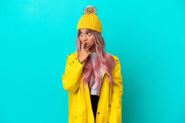 神経質で怖い青い背景で隔離の防雨コートを着ているピンクの髪の若い女性