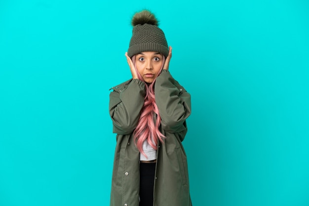 Young woman with pink hair wearing a rainproof coat isolated on blue background frustrated and covering ears