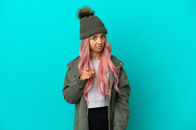 Young woman with pink hair wearing a rainproof coat isolated on blue background frustrated and pointing to the front