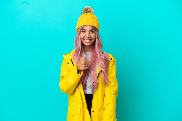 Foto giovane donna con i capelli rosa che indossa un cappotto antipioggia isolato su sfondo blu che fa un gesto di pollice in alto