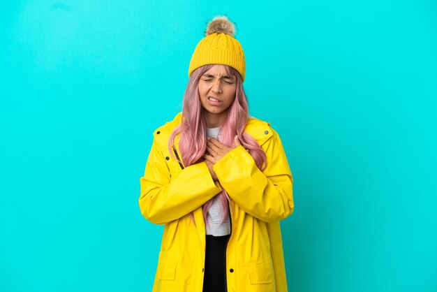 Young woman with pink hair wearing a rainproof coat isolated on blue background having a pain in the heart