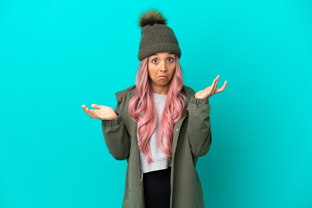 Young woman with pink hair wearing a rainproof coat isolated on blue background making doubts gesture