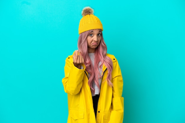 Young woman with pink hair wearing a rainproof coat isolated on blue background making money gesture
