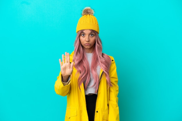 Young woman with pink hair wearing a rainproof coat isolated on blue background making stop gesture