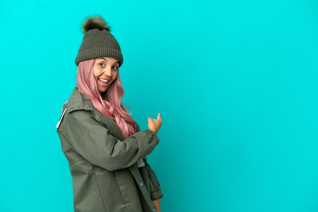 Young woman with pink hair wearing a rainproof coat isolated on blue background pointing back