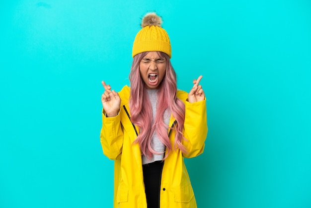 Young woman with pink hair wearing a rainproof coat isolated on blue background with fingers crossing