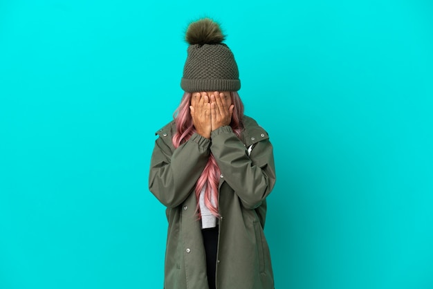 Young woman with pink hair wearing a rainproof coat isolated on blue background with tired and sick expression