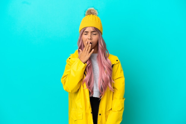 Young woman with pink hair wearing a rainproof coat isolated on blue background yawning and covering wide open mouth with hand