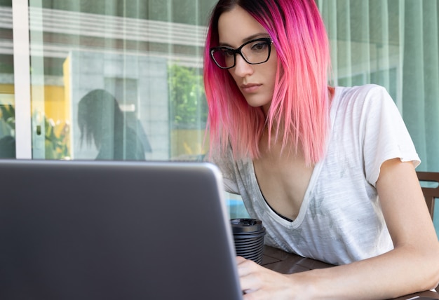 Молодая женщина с розовыми волосами работает с ноутбуком в кафе или домашней террасе