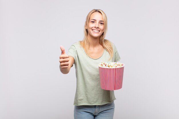 Young woman with a pop corns bucket feeling proud, carefree, confident and happy, smiling positively with thumbs up