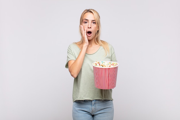 young woman with a pop corns bucket feeling shocked and scared, looking terrified with open mouth and hands on cheeks