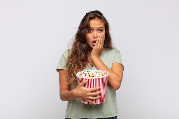 Young woman with a popcorn bucket feeling shocked and scared, looking terrified with open mouth and hands on cheeks