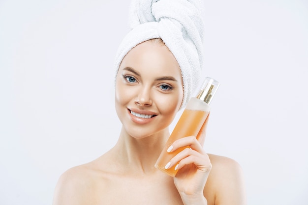 Young woman with positive smile, holds big bottle of aromatic perfume with flower odor