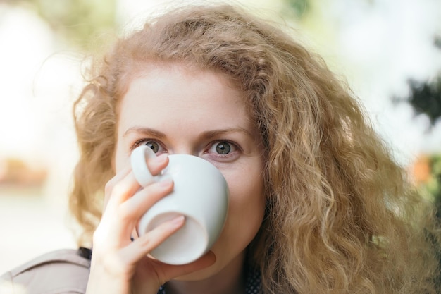 예쁜 얼굴과 곱슬곱슬한 긴 머리를 가진 젊은 여성이 야외에서 흰색 커피나 차 컵을 마시고 있다