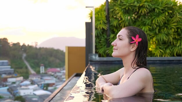 Young woman with purple flower in wet hair admires hilly landscape and blurry city buildings standing at hotel swimming pool edge