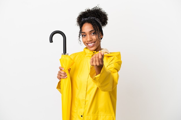 Young woman with rainproof coat and umbrella isolated on white background making money gesture