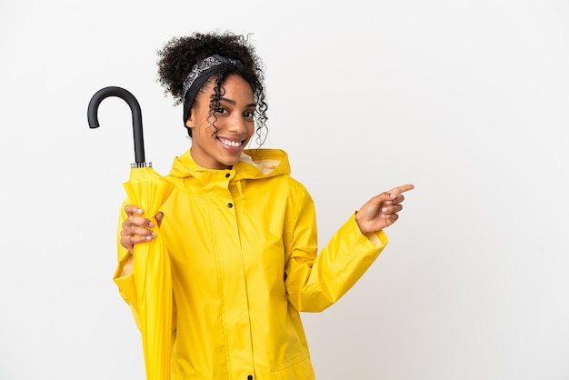 Young woman with rainproof coat and umbrella isolated on white background pointing finger to the side