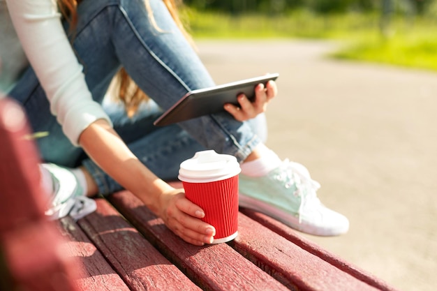 Молодая женщина с красным одноразовым бумажным стаканчиком кофе с помощью планшетного ПК сидит на красной скамейке в парке