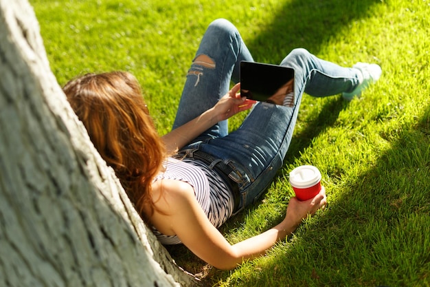 Young woman with red disposable paper cup of coffee using tablet pc under the tree on a green lawn in a park