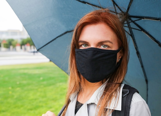 a young woman with red hair under a blue umbrella wearing a black protective mask