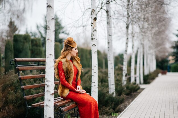 Young woman with red hair in a bright red dress on a bench in an empty park among the birches