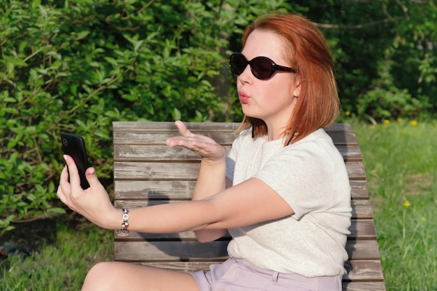 Young woman with red hair in sunglasses blows a kiss at the screen of his smartphone on his outstretched hand to take a photo of herself on phone. Woman makes a selfie on a smartphone in a city park.