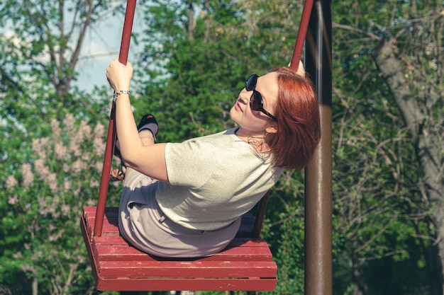 サングラスをかけた赤い髪の若い女性は、都市公園でブランコに揺れる笑顔。ブランコに乗っている女性は動きながら振り返った。大人のための子供向けエンターテインメント