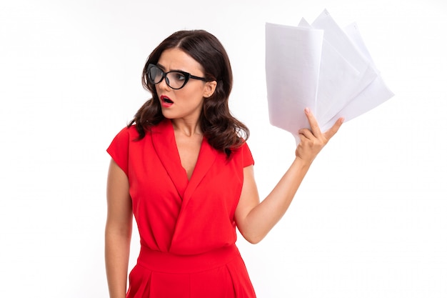 A young woman with red lips, bright makeup, dark wavy long hair, in a red suit, black glasses with transparent stacks stands, holds papers and is surprised