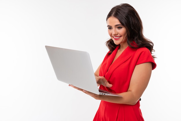 A young woman with red lips, bright makeup, dark wavy long hair, in a red suit, stands with a white laptop