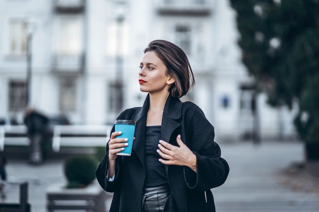 Молодая женщина с красными губами и короткой стрижкой, одетая в черное, позирует, держа в руках чашку кофе. За размытым городским фоном.