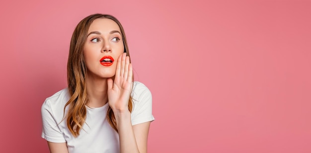 Young woman with red lipstick holds hand to face and whispers isolated over pink background