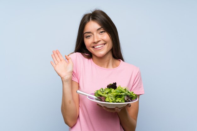 Young woman with salad over isolated blue background saluting with hand with happy expression