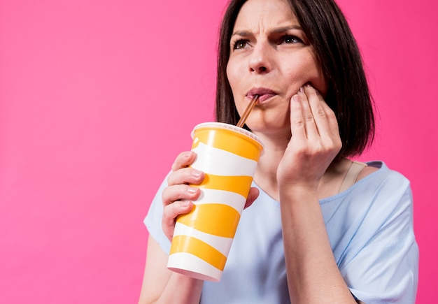 Young woman with sensitive teeth drinking cold water on color background